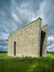 A photo of a contemporary private chapel in Suffolk, designed by Whitworth. In the background is a moody, cloudy sky. The chapel is surrounded by lush green grass.